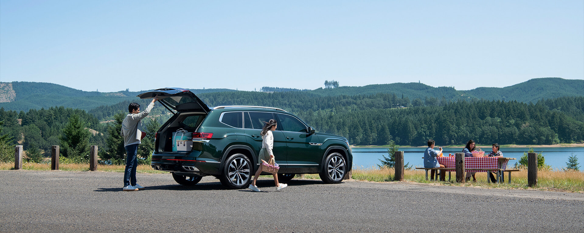 Person closing trunk of green 2023 Volkswagen atlas while family eats at picnic table near lake