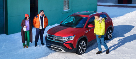 People standing around red 2023 Volkswagen Taos parked on snowy road next to turquoise building