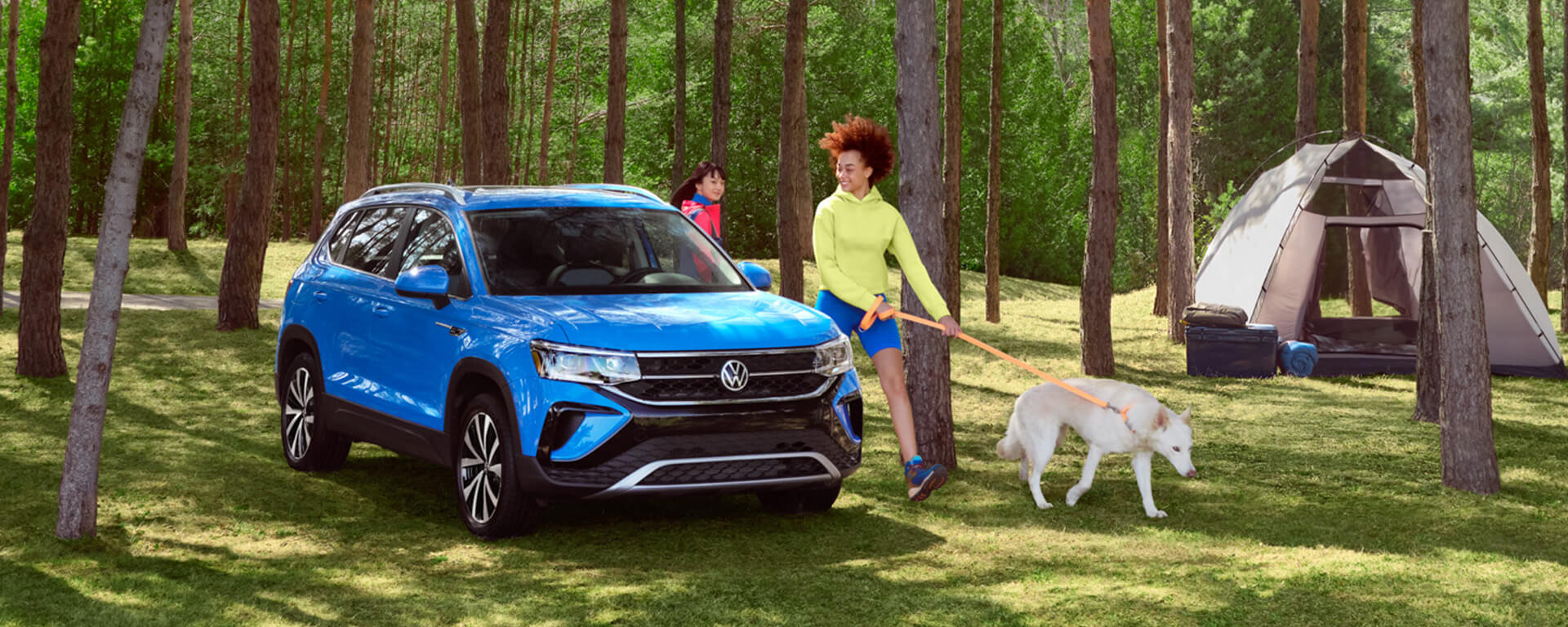 People with dog walking by blue 2023 volkswagen Taos parked in forest near camping tent