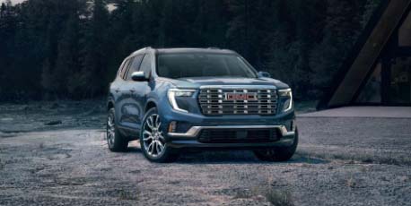 2025_gmc_acadia_suv_in_blue_parked_on_gravel_road_in_front_of_forest_near_modern_cabin_at_dusk