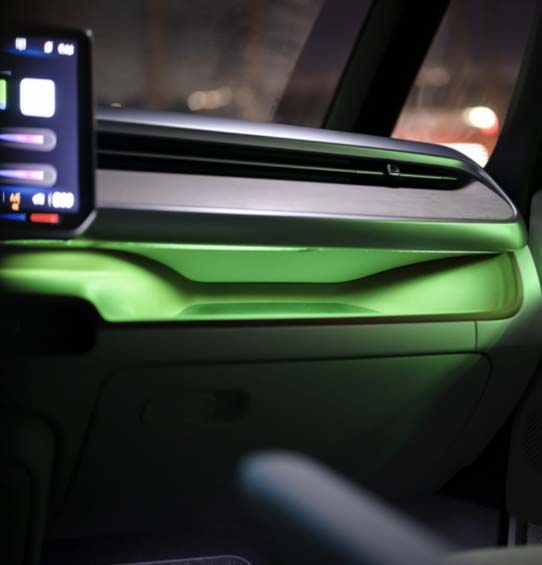 A close-up view of the dashboard in the Volkswagen ID. Buzz 2025, showcasing the customizable 30-color ambient lighting illuminating the passenger side, casting a vibrant green glow.