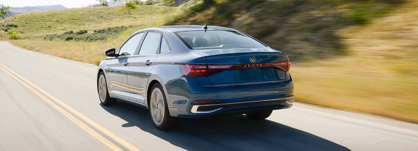 Rear view of the blue Jetta driving on a scenic road with a large tree and grassy hills in the background.