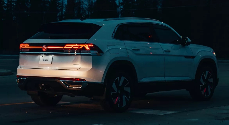 A white Atlas Cross Sport is parked on a forest road at dusk, with its rear lights illuminated.